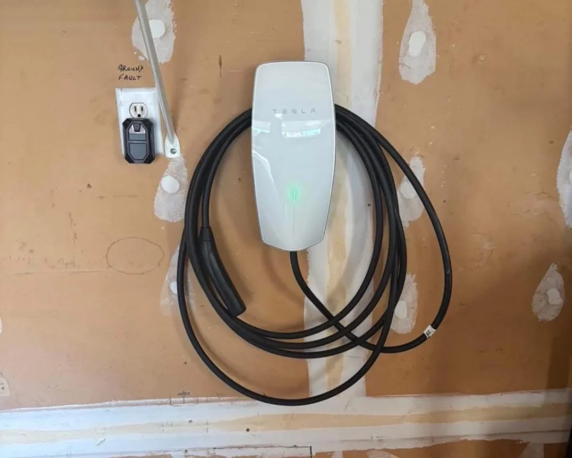 Tesla Wall Connector mounted on drywall with coiled black charging cable and standard outlet