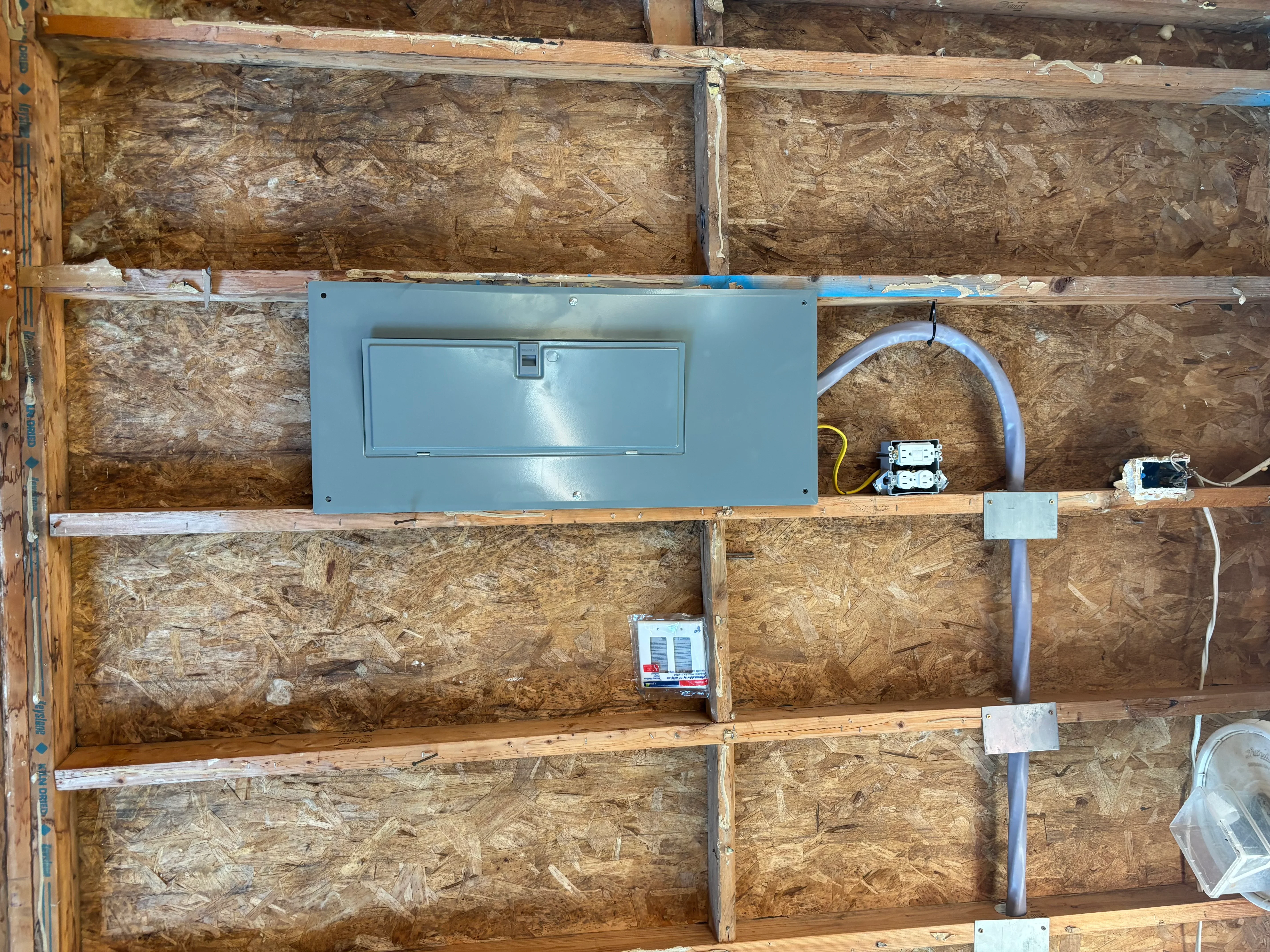 Blue electrical panel with circuit breakers, conduit, and outlet boxes installed in framed wall cavity with OSB sheathing